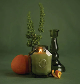 Lit candle in a green glass octagonal jar with embossed logo on the side is flanked by a green glass taper holder, orange and orange peel, and green plant sprig