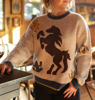Smiling model wears a beige sweater with blue and yellow scalloped edges and a central brown bucking bronco graphic accented by horseshoes, cactus, and sun symbols