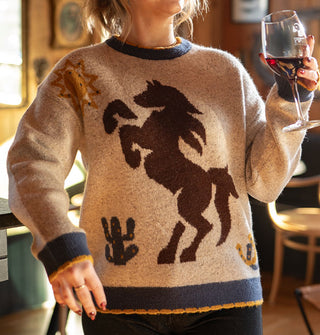 Model holding a wine glass wears a beige sweater with blue and yellow scalloped edges and a central brown bucking bronco graphic accented by horseshoes, cactus, and sun symbols