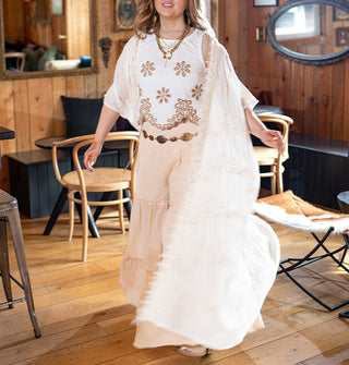 Model wears a long white kimono with subtle floral embroidery and fringe paired with a floral embroidered crop top and tiered wide leg pants