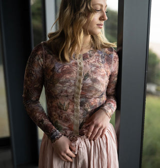 Model gazing out a window wears a long-sleeved floral print top with snap button front and tan placket