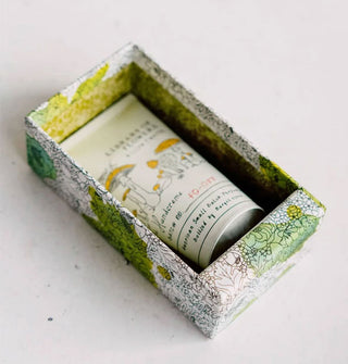 A tube of Library of Flowers Willow & Water Handcreme rest inside a decorative floral-patterned gift box with green and chartreuse spot-coloring