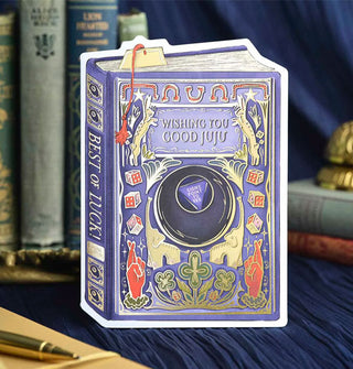 Greeting card is designed to resemble a book with a central Magic 8 Ball design in which the fortune printed is, "Signs point to yes," under the title, "Wishing You Good Juju," all surrounded by good luck symbols, and a message on the spine that reads, "Best of luck"