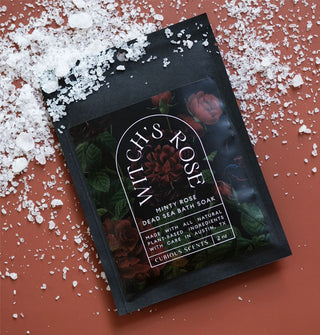 Black packet of Witch's Rose Minty Rose Dead Sea Bath Soak with dark rose motif and white lettering rests on a terracotta-colored surface scattered with coarse salt