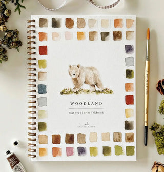 Spiral-bound Woodland Watercolor Workbook featuring paint swatch designs as a border around a central bear artwork is staged with paint tube, paintbrush, ribbon, and miscellaneous plants