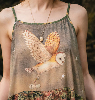 Model wears a sleeveless dress with illustration of an owl in flight carrying a scroll in its talons on the bodice