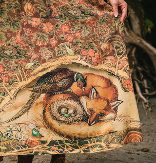Skirt with illustration of a fox curled around a pheasant's nest of eggs, the pheasant perched on the fox's hind leg, under a branch that serves as the border to a densely floral printed backdrop print