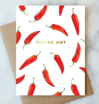 White greeting card featuring artwork of red chili peppers with green stems rests on top of a kraft paper envelope and says, "You're hot" in gold foil lettering