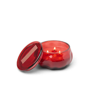 Red glass jar candle with three lit wicks inside and a matching lid propped up alongside